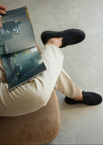 Person wearing black shoes and beige pants sitting on a couch with a book on their lap.
