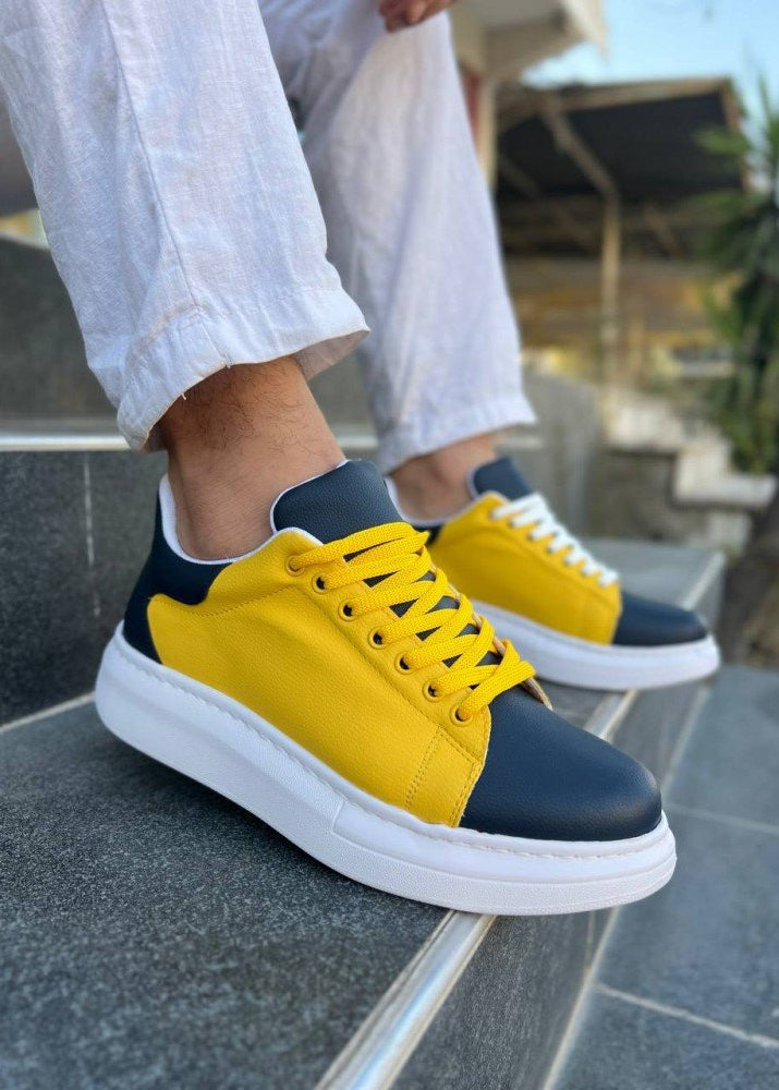 Yellow and navy sneakers worn with white pants on a stone surface.