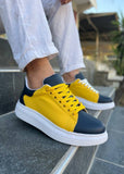 Yellow and navy sneakers worn with white pants on a stone surface.