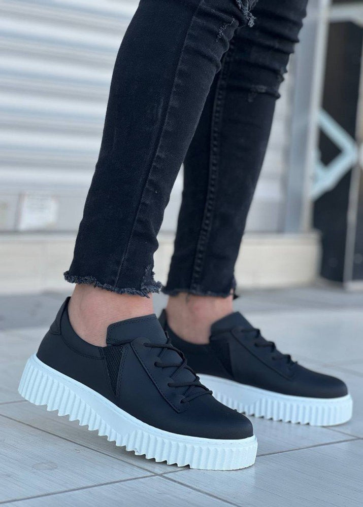 Black sneakers with white soles worn by a person on a tiled floor.