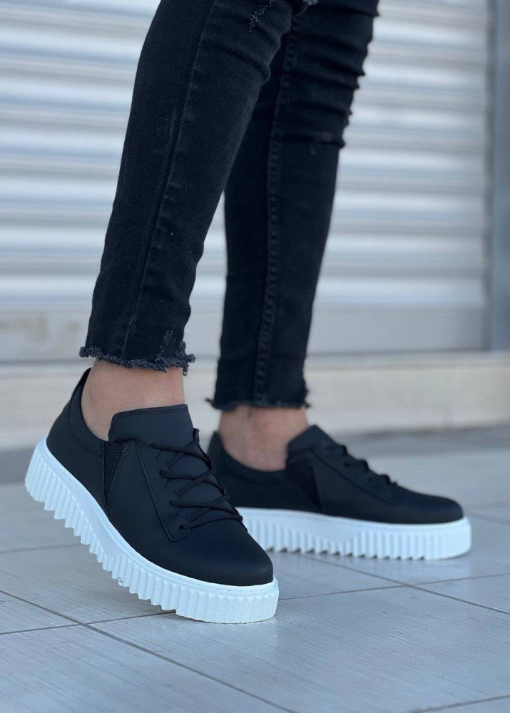 Black sneakers with white soles worn by a person on a tiled floor.